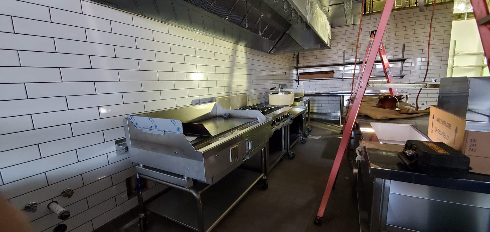 Restaurant kitchen with a griddle and stainless equipment being set under a hood, white tile walls