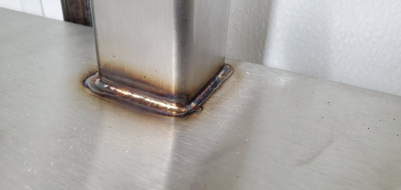 Close-up of a stainless steel weld where a square table leg meets the top, bead ground flat
